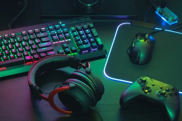 A close-up shot of gaming peripherals including a mechanical keyboard with glowing RGB keys, a headset, a gaming mouse on a mousepad, and a black game controller, all lit with green and magenta light.